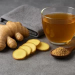 قطع من الزنجبيل الطازج مع شرائح وجذور كاملة، وشاي زنجبيل ساخن في كوب زجاجي، وملعقة خشبية تحتوي على مسحوق زنجبيل على طاولة رمادية.